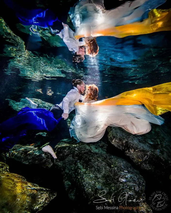 Underwater photographer in Playa del Carmen capturing a Trash The Dress photoshoot in a cenote, bride and groom kissing on the rocks surrounded by flowing yellow and blue fabric with their reflection on the water surface.