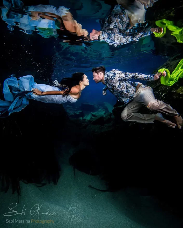 Underwater photographer in Playa del Carmen capturing a Trash The Dress photoshoot in a cenote, bride in a white dress swimming toward her groom holding bright green fabric, with their reflection on the water surface.
