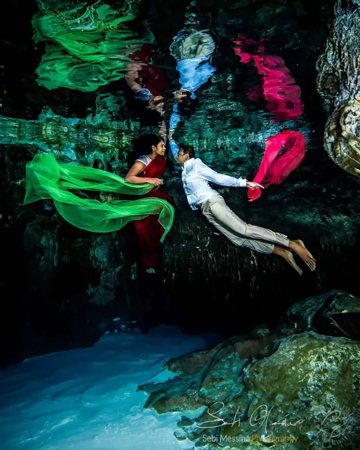 Underwater photographer in Playa del Carmen capturing an Indian couple in a cenote Trash The Dress session, bride in a red sari with flowing green fabric and groom in a white shirt holding pink fabric, both reflected on the water surface.