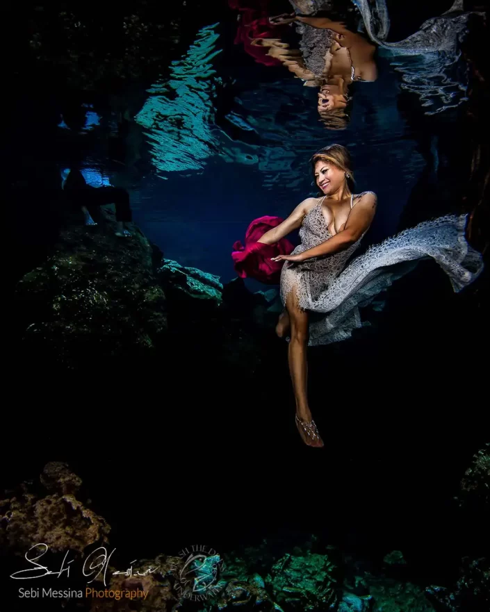 Underwater photographer in Playa del Carmen capturing a woman in a lace dress during an underwater modeling Trash The Dress session in a cenote, holding red fabric with her reflection on the water surface.