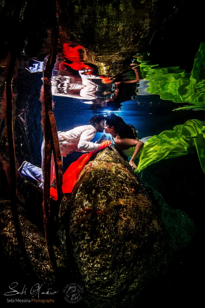 Underwater engagement photoshoot in a Riviera Maya cenote between Playa del Carmen and Tulum, Mexico: couple kissing beside a limestone ledge with surface reflections and colorful fabrics.