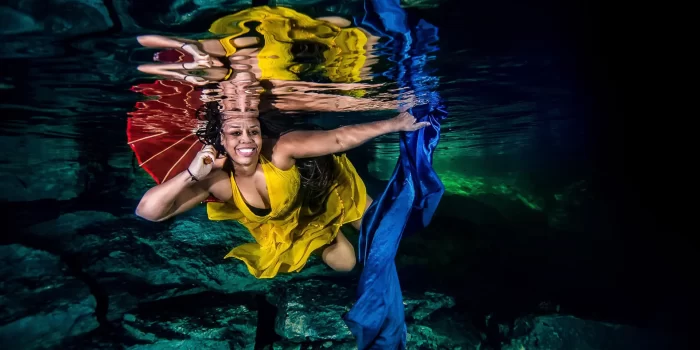 Underwater modeling photoshoot in a Riviera Maya cenote near Tulum: smiling woman in a yellow dress swims beneath the surface holding a red parasol while deep-blue fabric trails behind, with mirror-like reflections above.