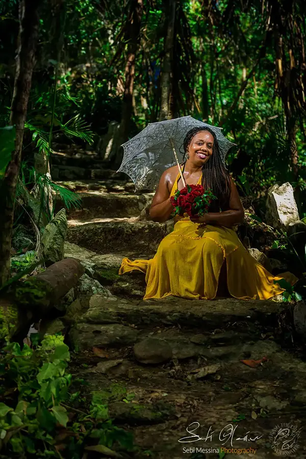 Woman in a yellow dress sits on stone steps in the jungle near a cenote close to Tulum, holding a lace parasol and a bouquet of red roses.