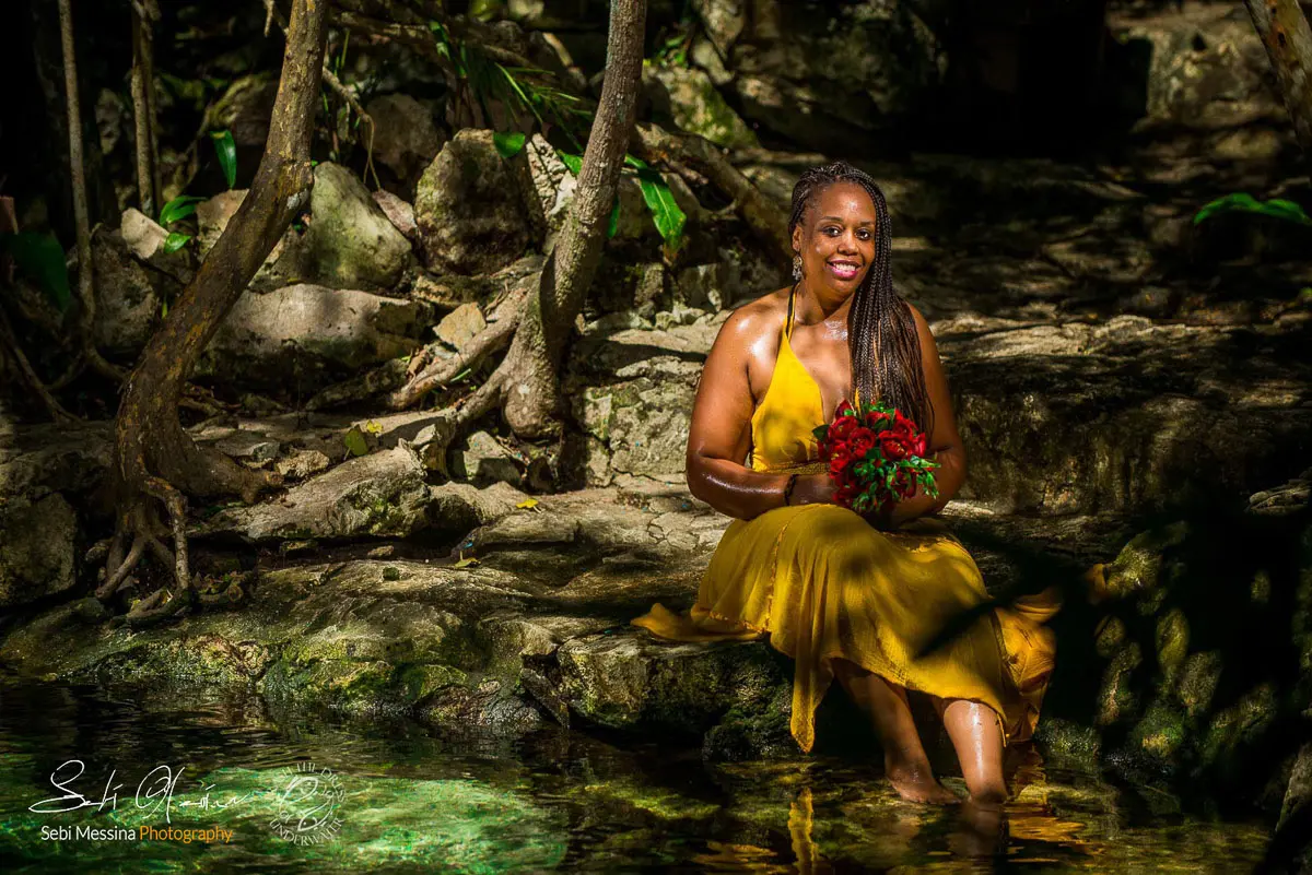 Cenote portrait near Playa del Carmen: woman in a yellow dress sits on rocks beside shallow crystal-clear water, holding a bouquet of red roses in dappled jungle shade.