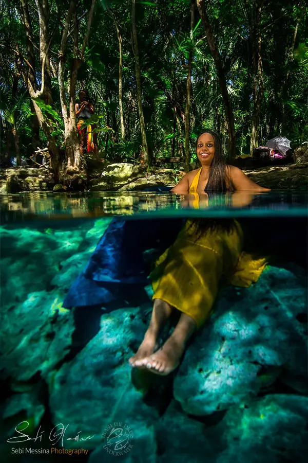 Over-under split shot in a cenote near Tulum: woman relaxes underwater on pale rocks in a flowing yellow dress while the jungle appears above the waterline.
