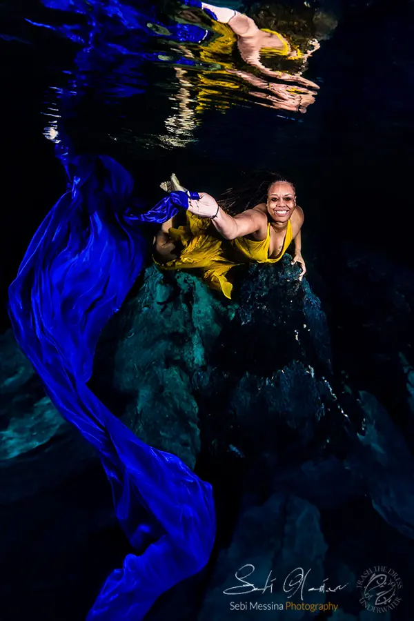 Underwater modeling in a cenote near Playa del Carmen: smiling woman in a yellow dress poses on dark rocks as long blue fabric streams through clear water, reflected on the surface.