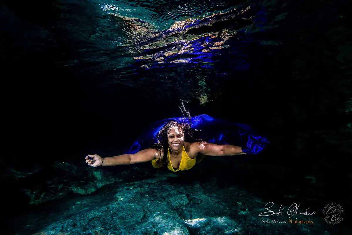 Underwater modeling photoshoot near Tulum: woman swims forward with arms extended over turquoise rock, draped in deep-blue fabric beneath a rippled water surface.