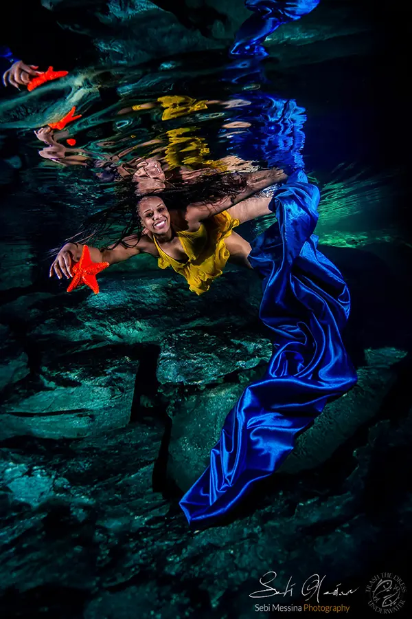 Underwater cenote photoshoot near Playa del Carmen: woman in a yellow dress holds two red starfish while blue fabric cascades downward, with reflections on the water surface.