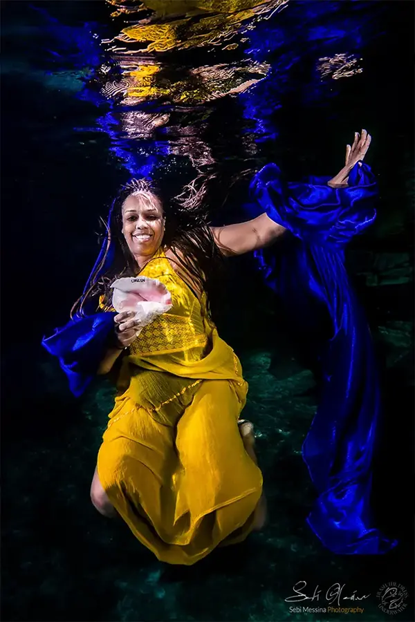 Underwater photoshoot in a cenote near Tulum: woman in a flowing yellow dress holds a large shell and smiles toward the camera while blue fabric curls beside her in dark, clear water.