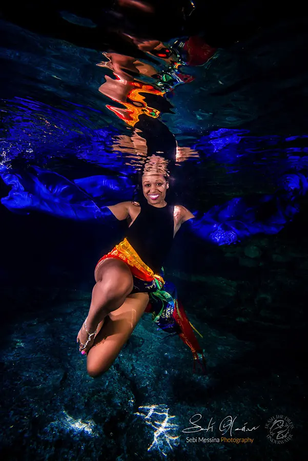 Underwater modeling scene in a cenote near Playa del Carmen: woman poses in a black swimsuit with a bright multicolor skirt as blue fabric billows out like wings beneath the shimmering surface.
