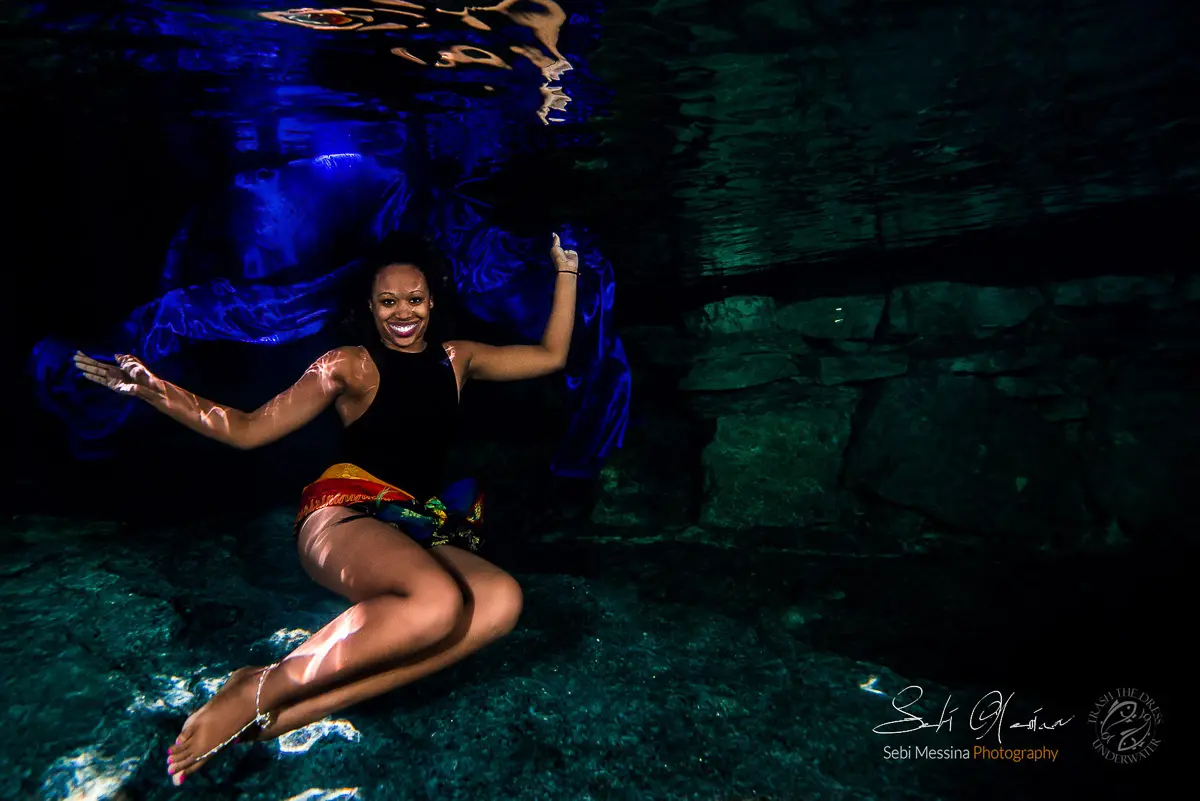 Underwater modeling photoshoot in a Riviera Maya cenote near Tulum: woman in a black swimsuit and colorful sarong sits in a relaxed pose over rocky ground while deep-blue fabric drifts behind her in dark, clear water.
