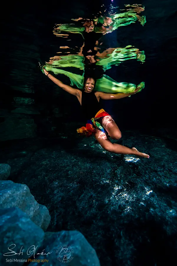 Underwater cenote photoshoot near Playa del Carmen: woman in a black swimsuit and bright sarong spreads flowing green fabric like wings, with a strong surface reflection above and textured limestone below.