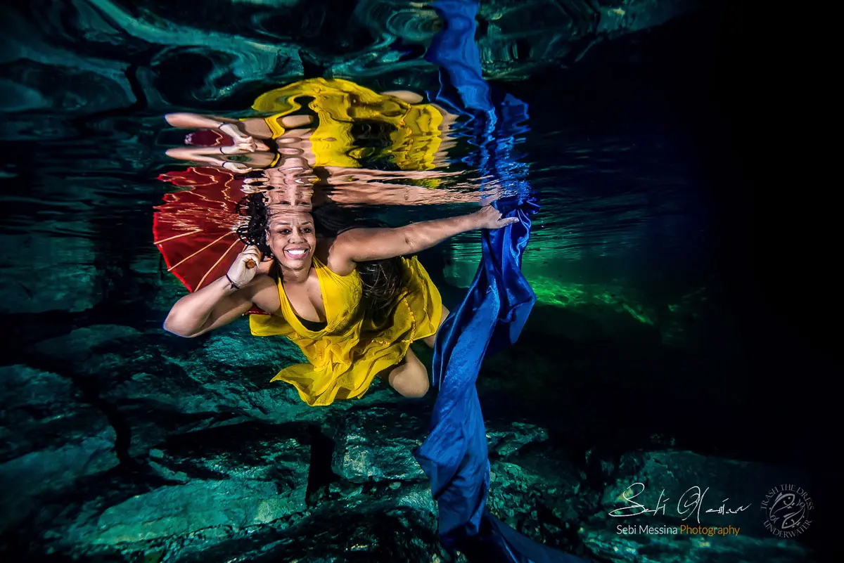 Underwater modeling in a cenote near Tulum: smiling woman in a yellow dress swims just beneath the surface holding a red parasol while a long blue fabric ribbon trails beside her, reflected in the rippled water.