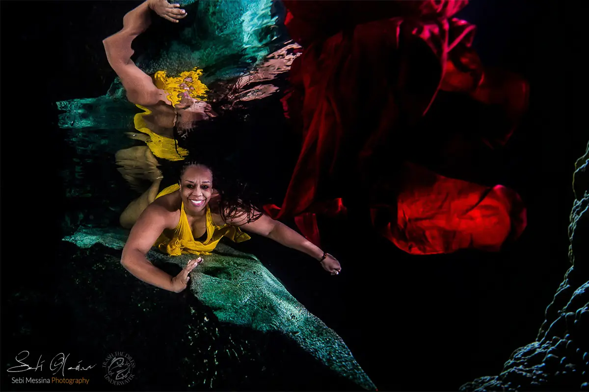 Underwater photoshoot in a Riviera Maya cenote near Playa del Carmen: woman in a yellow dress poses near a rock ledge while deep-red fabric billows in the dark water, creating a bold color contrast and surface reflection.
