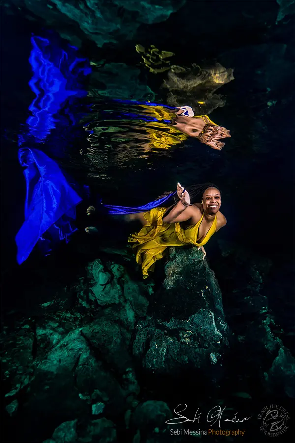 Underwater modeling photoshoot near Tulum: woman in a yellow dress reaches forward over rugged cenote rocks as blue fabric floats to the side; her reflection shimmers on the surface above.
