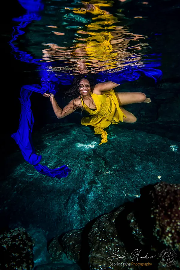 Underwater cenote photoshoot near Playa del Carmen: woman in a flowing yellow dress floats near the surface while she holds a long blue fabric ribbon that curves into a circle, with bright ripples and reflections overhead.