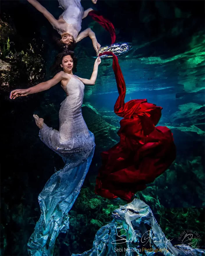 Underwater photographer in Playa del Carmen capturing a Trash The Dress portrait in a cenote, bride in a white lace dress floating with swirling red fabric and her reflection on the water surface.