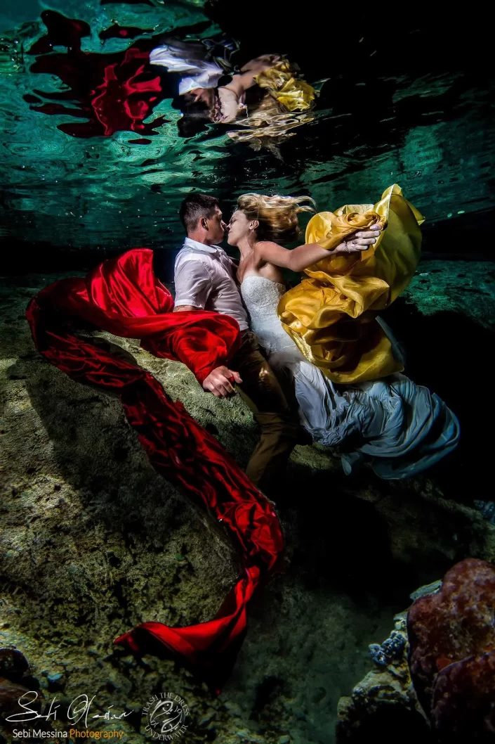 Bride and groom kissing underwater in Cenote El Buho, Riviera Maya, with dramatic red and gold fabrics floating in the scene.